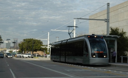 Having crossed intersection, Houston LRT train accesses station on Fannin St. as traffic control system allows queue of motor vehicles to make left turn over track reservation behind it.