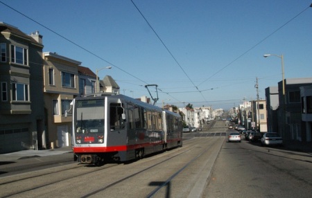 In this view of single-car train on slightly raised median near 16th Avenue, transverse spanwire that holds OCS power wire can be seen behind train, suspended between TES poles on either side of street. TES poles also serve as street light masts, a typical dual function. PHOTO: Peter Ehrlich.